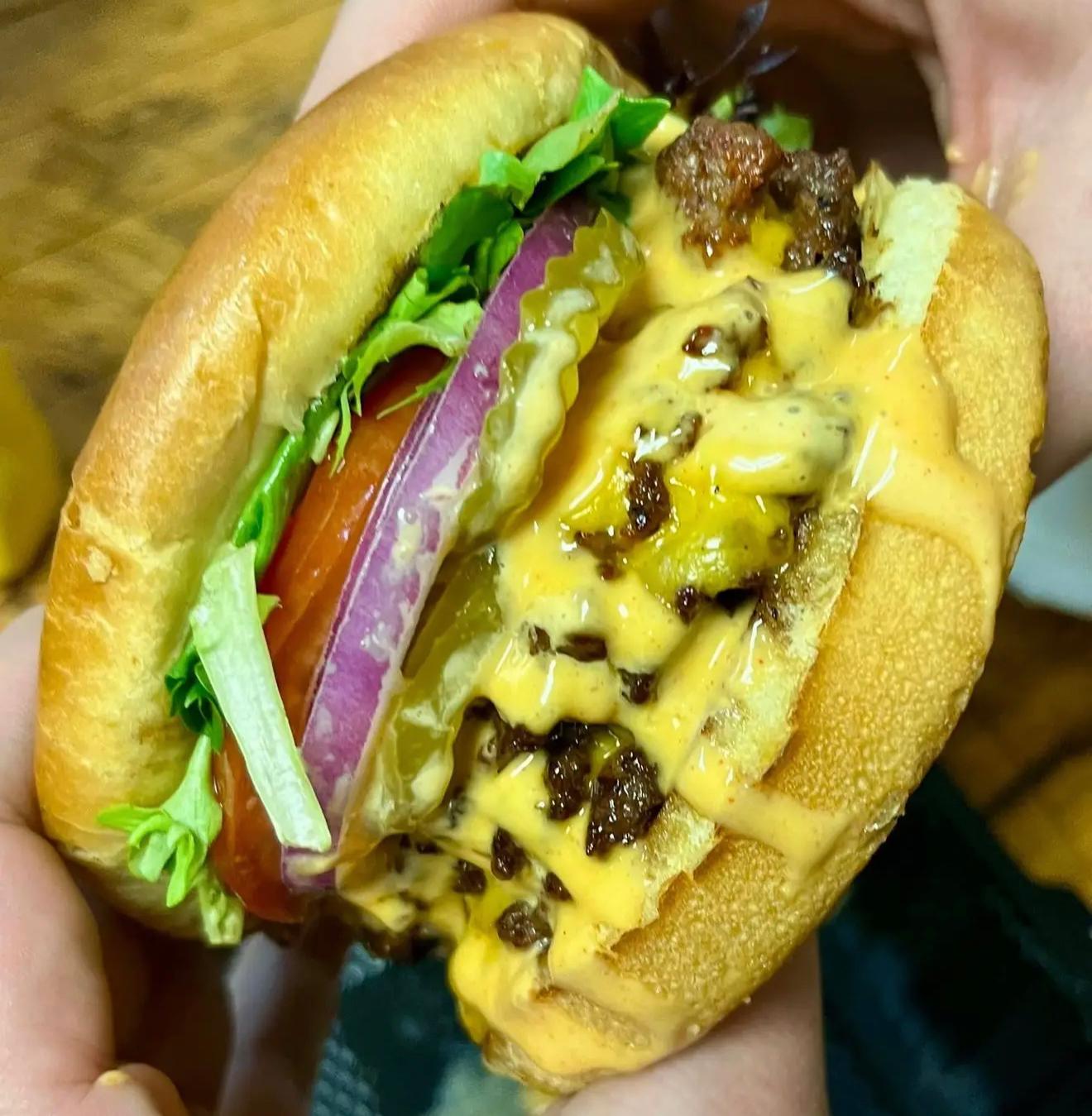 The BurgerBurger held in hand — a double smash patty with melted American cheese, lettuce, tomato, red onion and pickle on a toasted bun